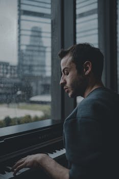 A thoughtful musician plays piano by a rainy window.
