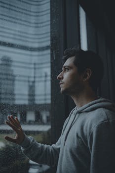 A man in a hoodie gazes out a rain-streaked window reflecting urban city vibes.