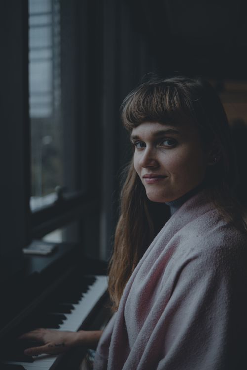Free Young woman playing piano by a window with a serene expression, wrapped in a blanket. Stock Photo