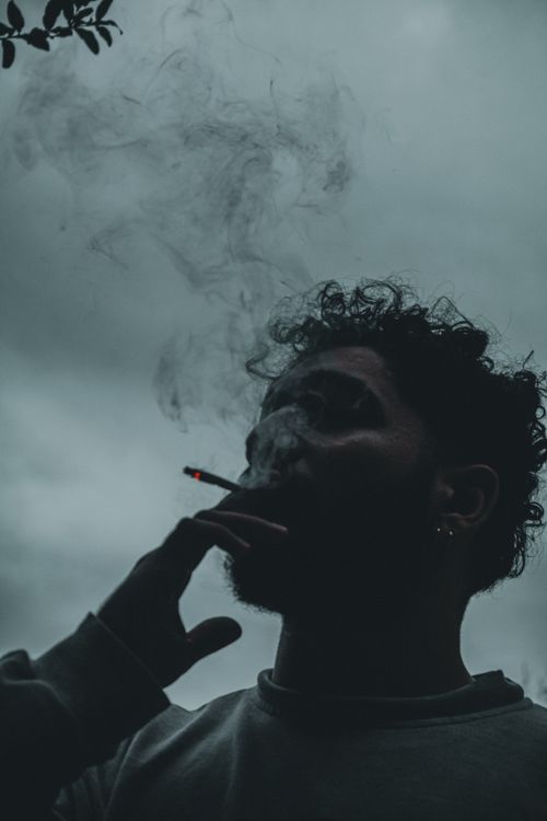 Free Moody silhouette of a bearded man smoking a cigarette with curling smoke against a cloudy sky. Stock Photo