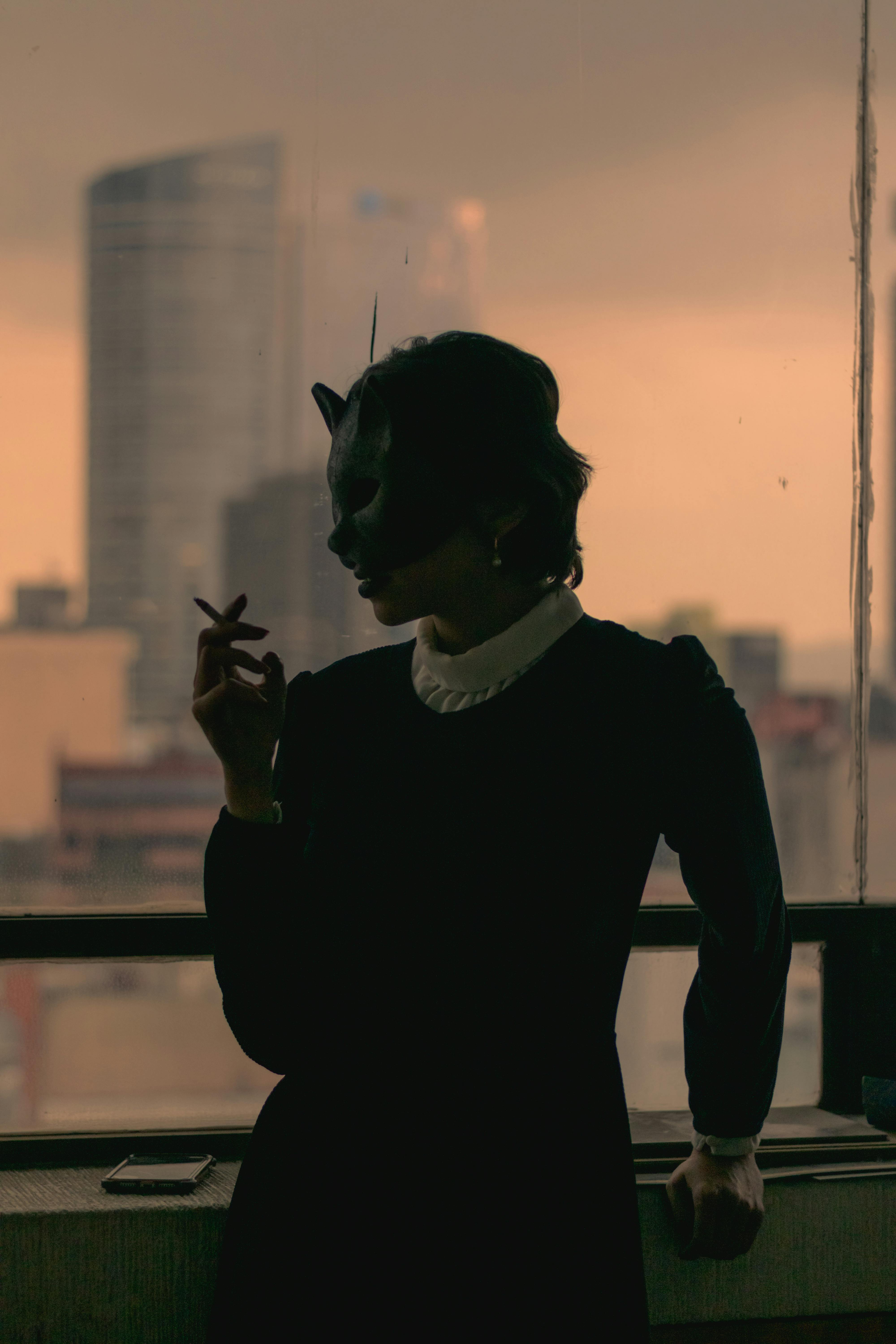 Silhouette of a stylish woman with a cat mask smoking indoors against an urban backdrop.