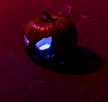 Close-up of a carved pumpkin with an eerie blue glow against a dark, red-lit background, perfect for Halloween themes.