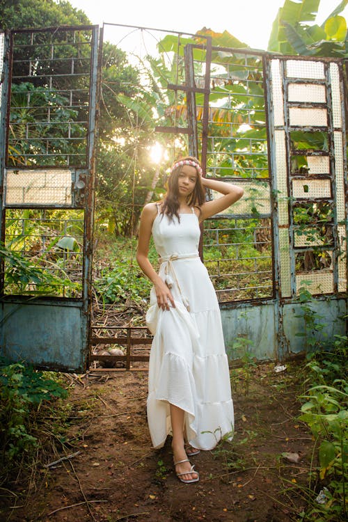 Woman in White Spaghetti Strap Dress Standing Beside a Metal Gate