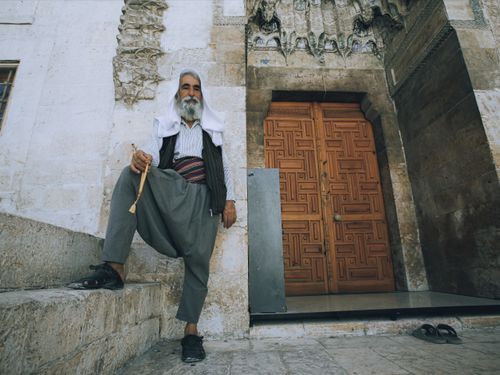Free An elderly man in traditional attire sits by a mosque entrance, embodying spirituality and culture. Stock Photo