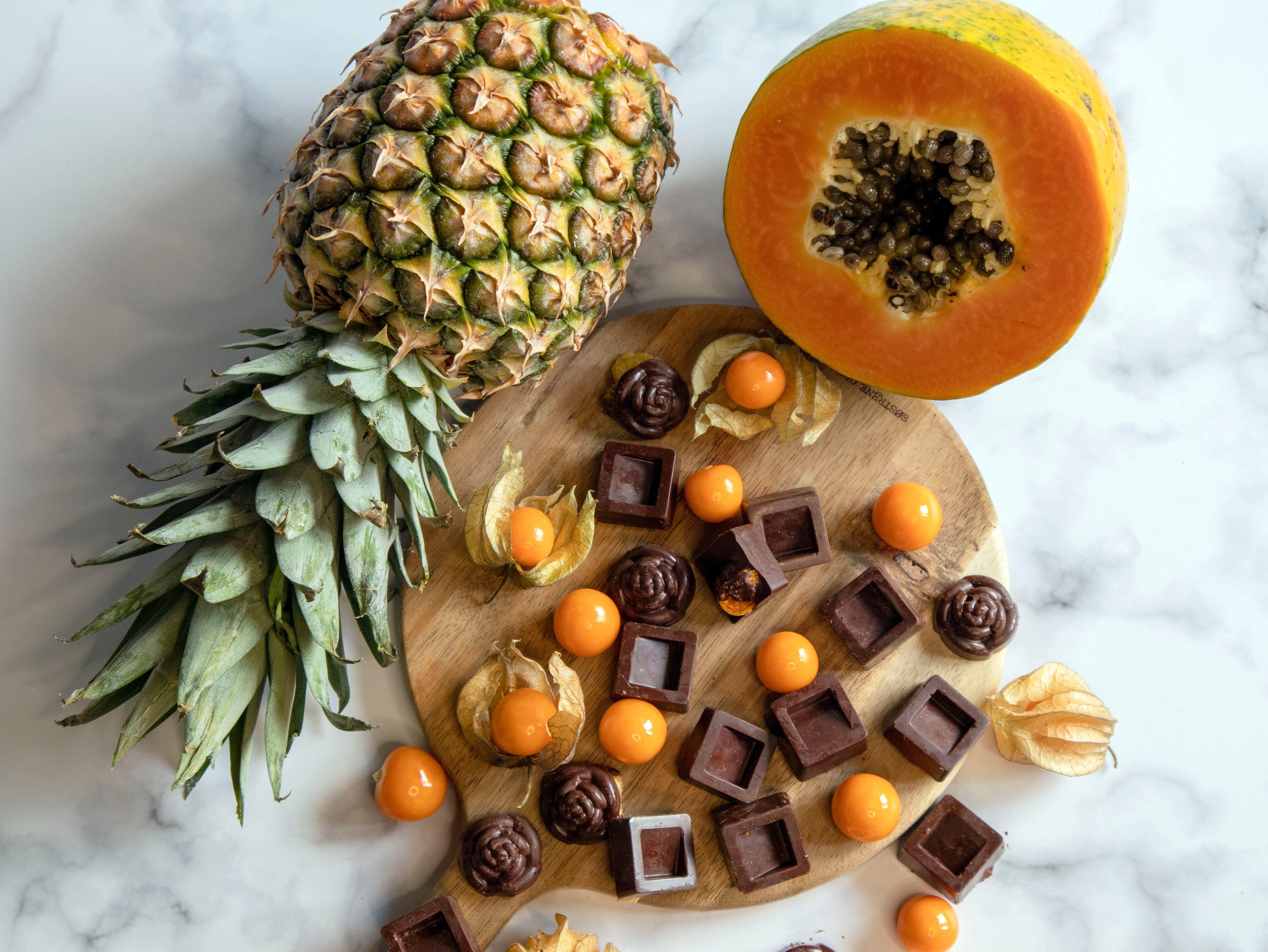 A vibrant arrangement of pineapple, papaya, and assorted chocolates on a wooden board and marble surface.