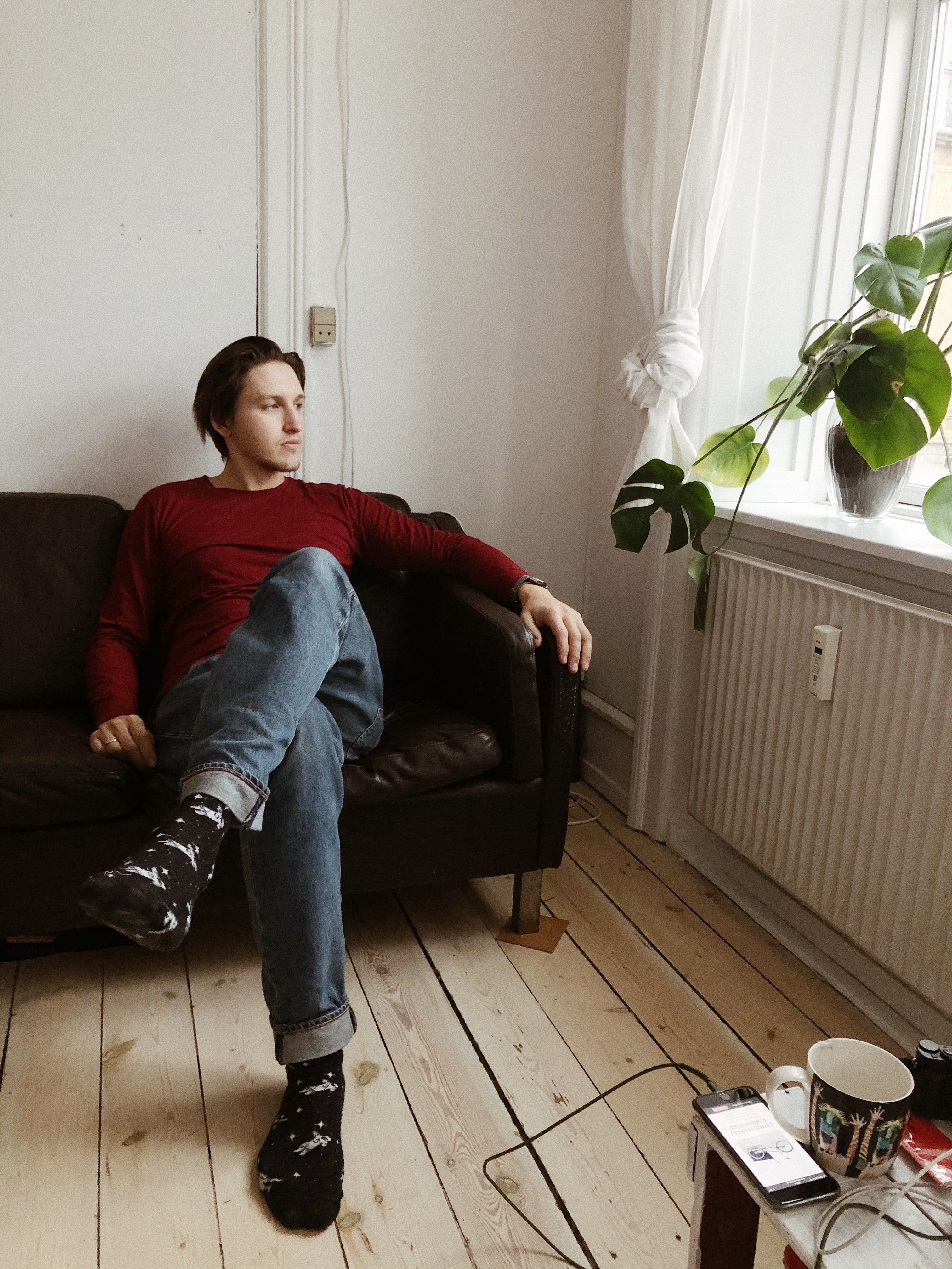 Casual scene of a man in a red sweater sitting on a sofa by a window with natural light.