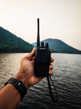 Close-up of a hand holding a walkie talkie with a lake and mountains in the background.