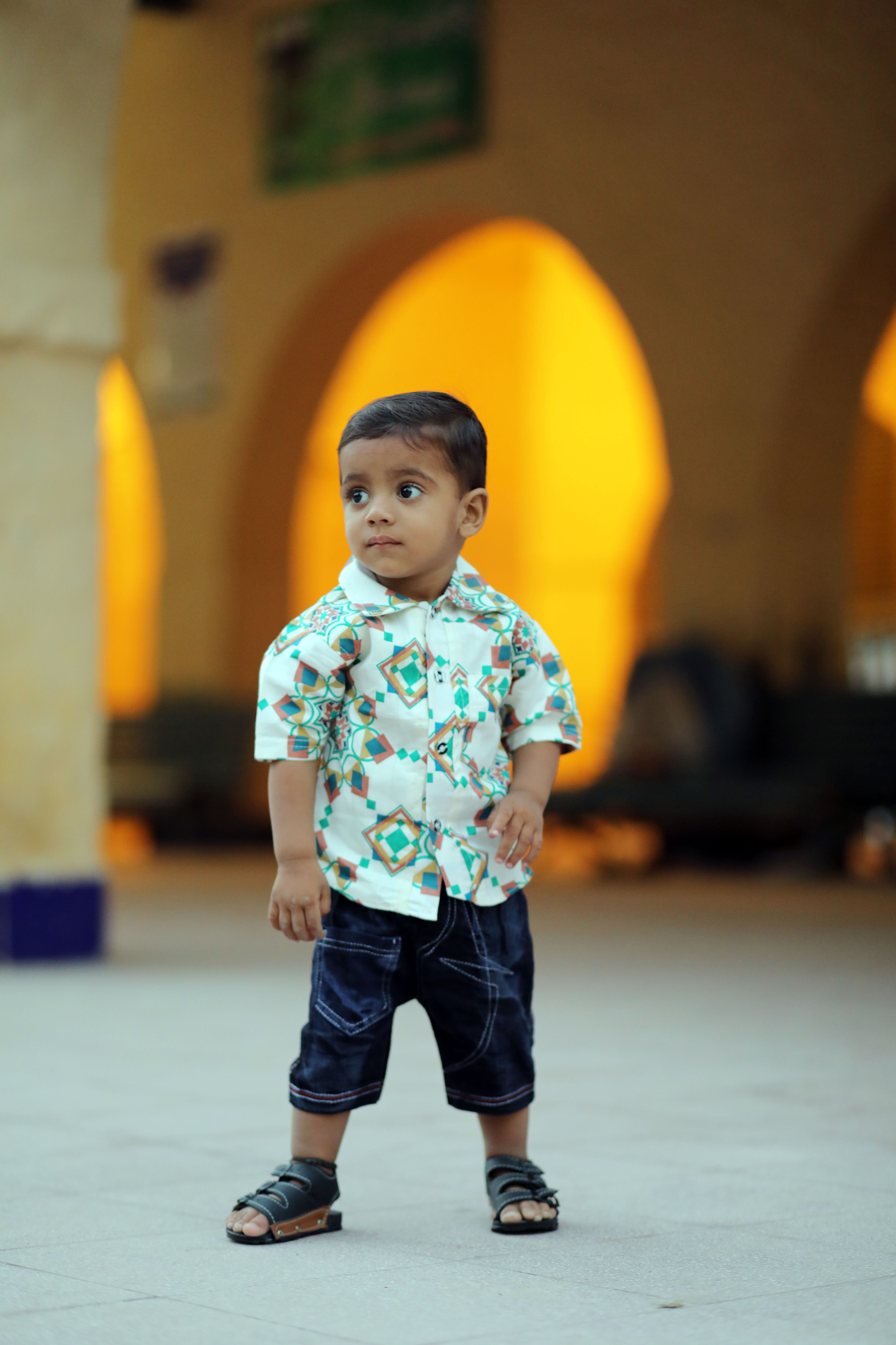 A young boy in vibrant clothing stands indoors, looking away, with warm lighting in the background.