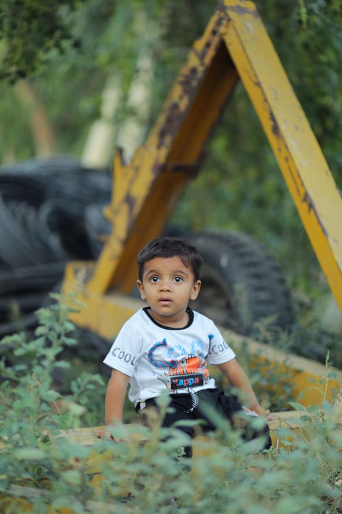 Free A young child sits amidst rusty machinery and lush plants in a playground. Stock Photo