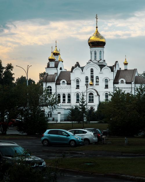 Free Beautiful view of Holy Trinity Orthodox Church in Kemerovo at dusk with golden domes and parked cars. Stock Photo