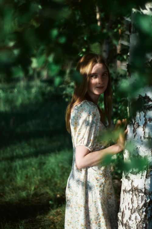 Free A redhead woman in a floral dress stands by birch trees in a lush green summer forest. Stock Photo