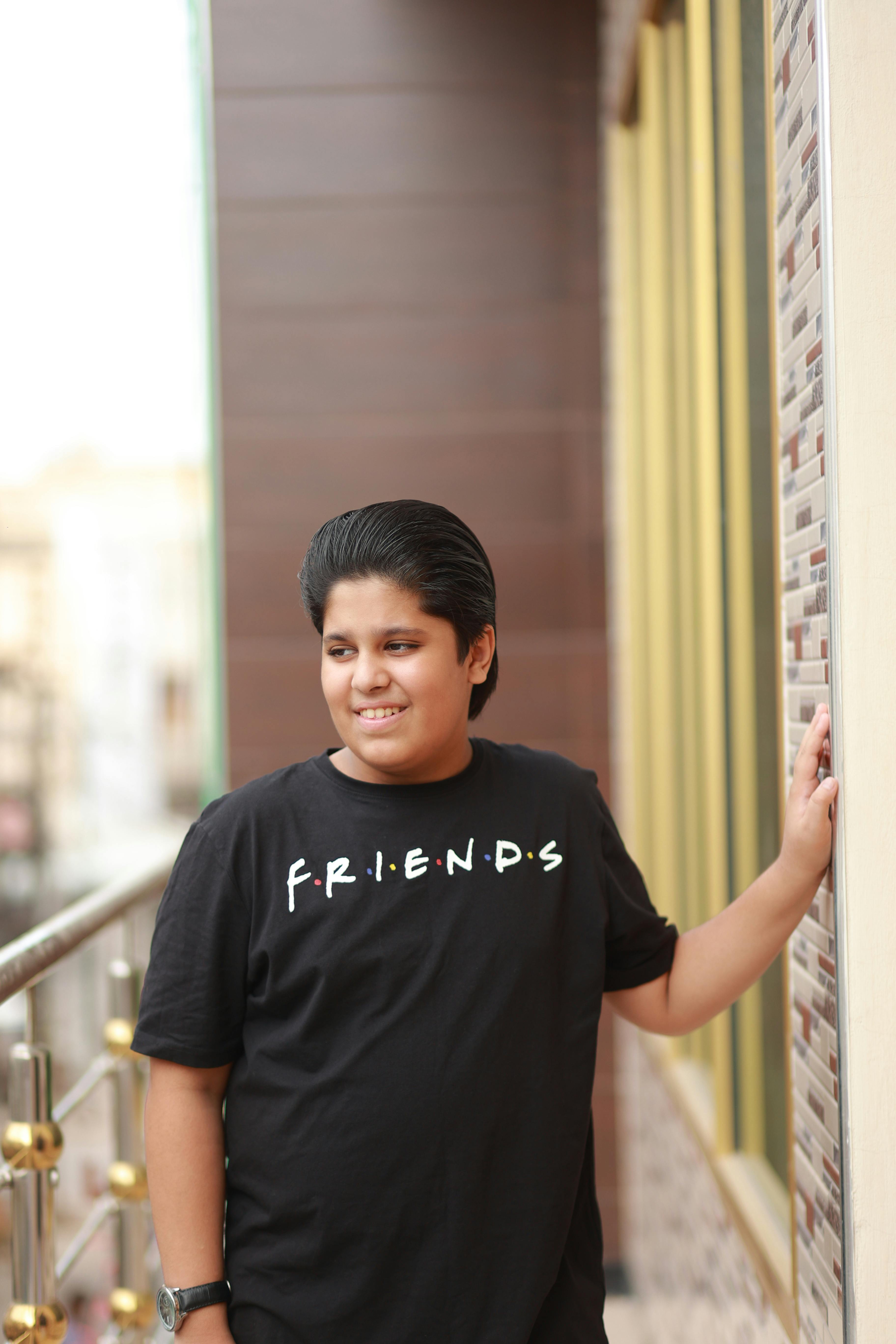 A cheerful teenager in a FRIENDS t-shirt smiles while leaning on a balcony in an urban setting.