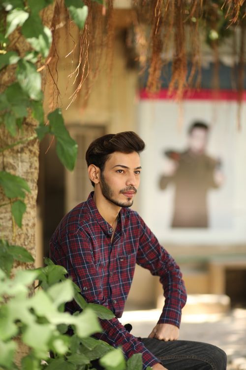 Free A man in a plaid shirt sits outdoors, surrounded by greenery on a sunny day. Stock Photo