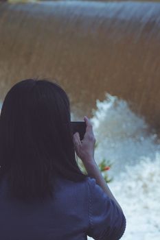 A woman capturing a waterfall scene on her smartphone in a serene outdoor setting.