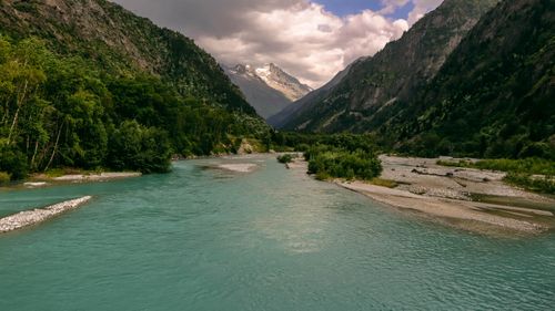 Free A tranquil river flowing through a lush valley surrounded by towering mountains under an overcast sky. Stock Photo