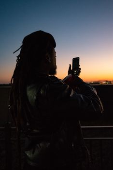 A man in a leather jacket captures a stunning sunset on his smartphone, silhouetted against the fading light.