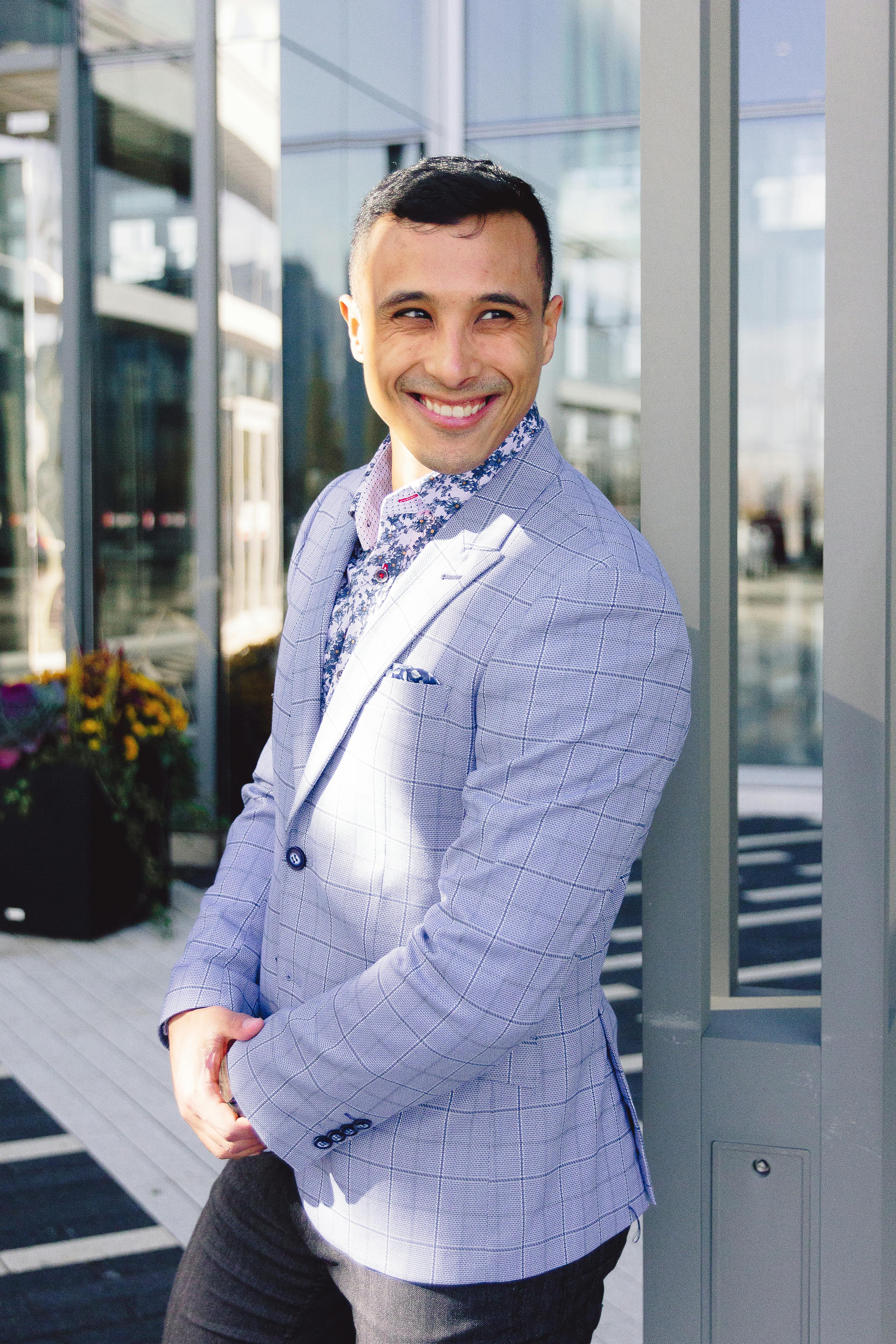 A young man joyfully smiling in a light checkered suit, standing outdoors.