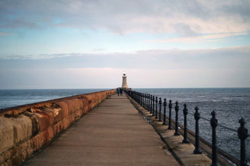 Free A long pier extending to a distant lighthouse under a calm sky and ocean backdrop. Stock Photo