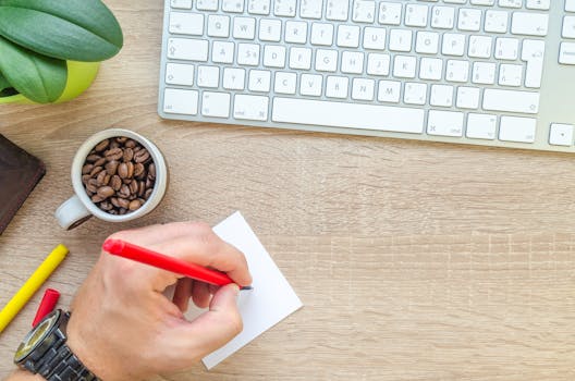 A workspace featuring a keyboard, coffee beans, note, and a hand writing with a red pen.