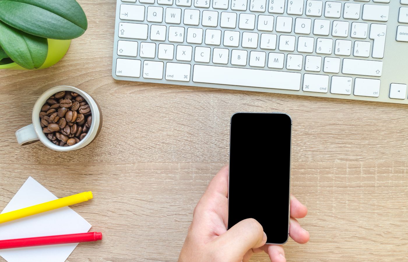 Overhead view of a hand holding smartphone near keyboard, mug of coffee beans, and plant on wooden desk.