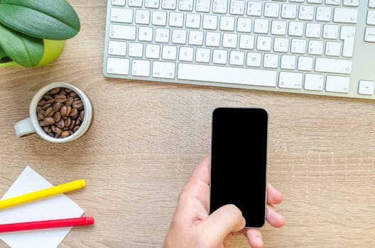 Overhead view of a hand holding smartphone near keyboard, mug of coffee beans, and plant on wooden desk.