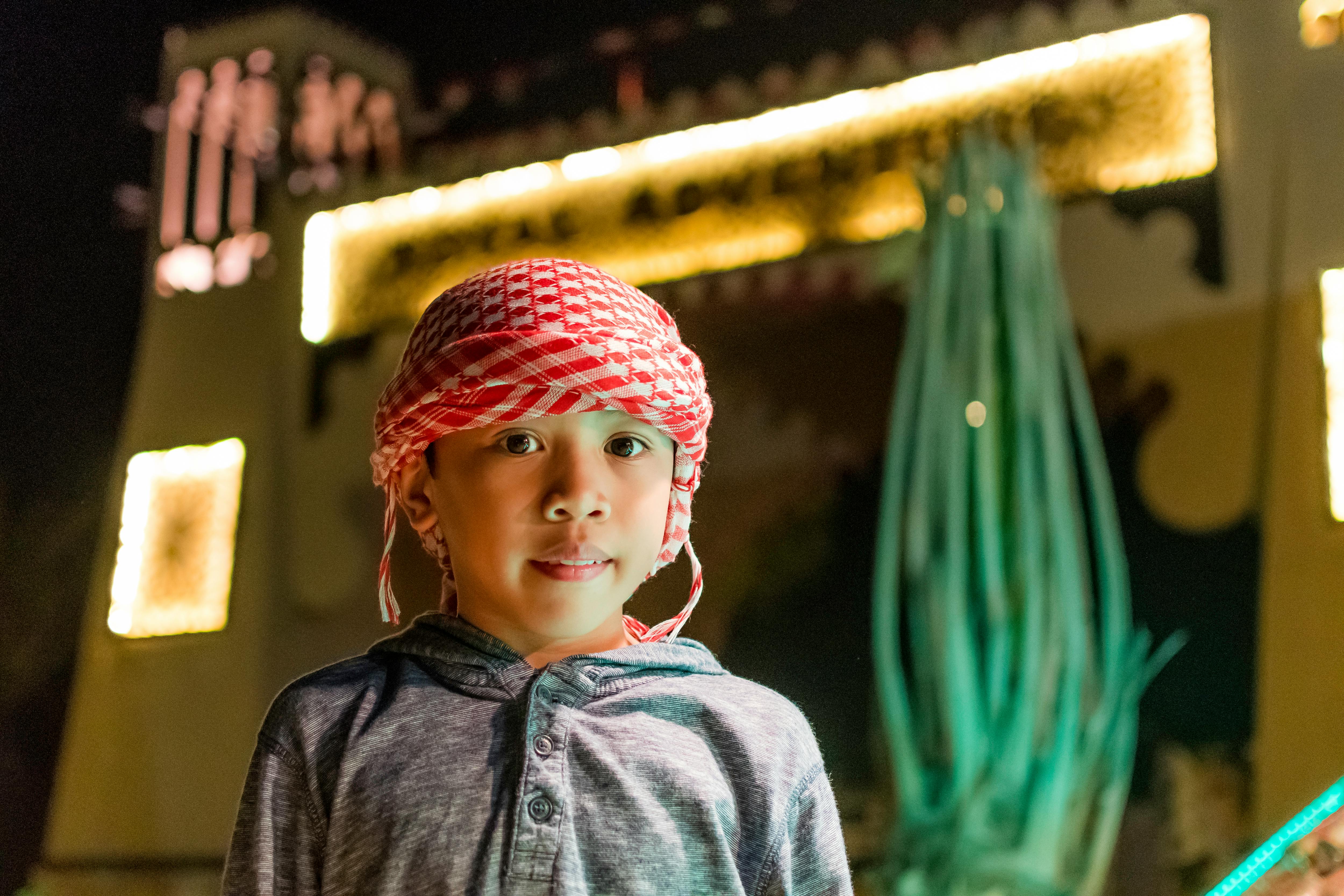 A young boy in traditional headwear stands brightly lit against a festival backdrop at night.