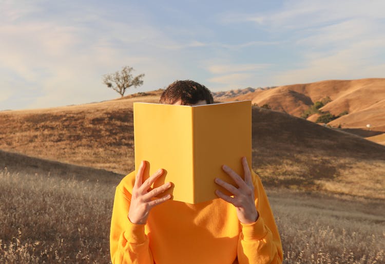 Teenage Boy Hiding Behind A Yellow Book In A Landscape