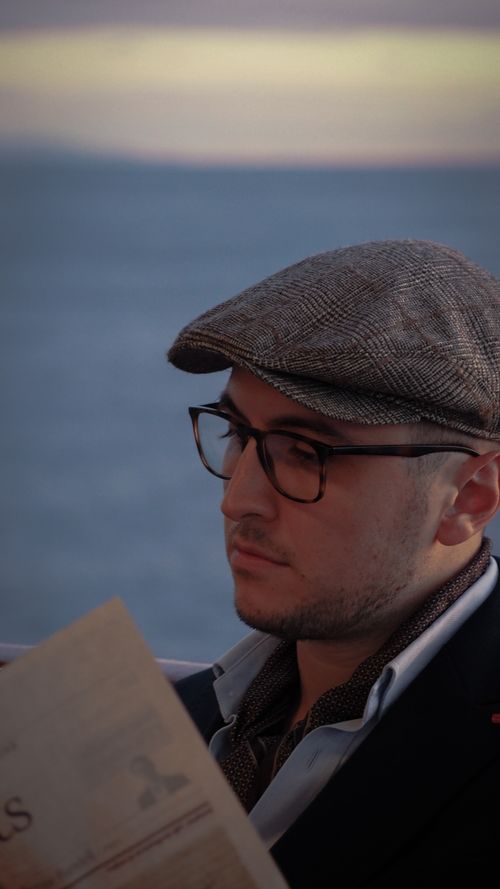 Free A man with eyeglasses and a flat cap reading a newspaper by the ocean during sunset. Stock Photo