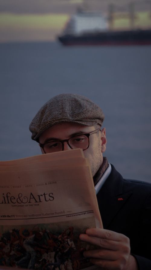 Free A man wearing a flat cap and eyeglasses reads a newspaper outdoors by the water. Stock Photo