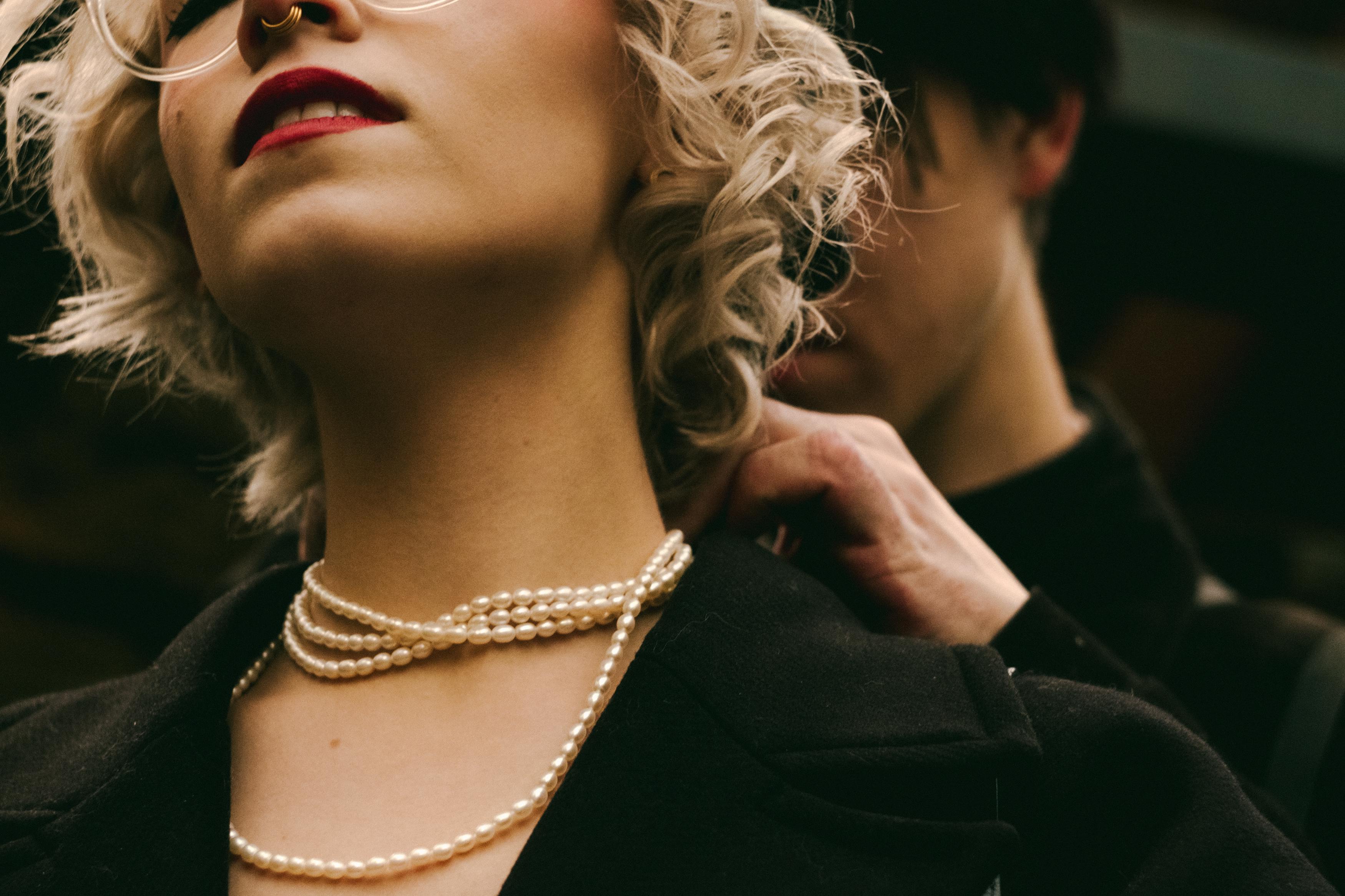 An elegant woman with red lipstick wearing a pearl necklace as someone helps her fasten it.
