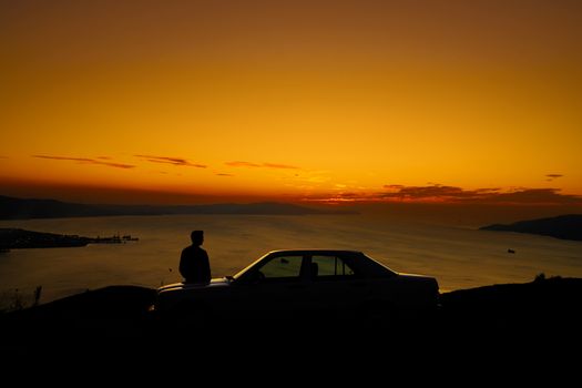 Dramatic silhouette of a man beside a parked car with a breathtaking ocean sunset view.