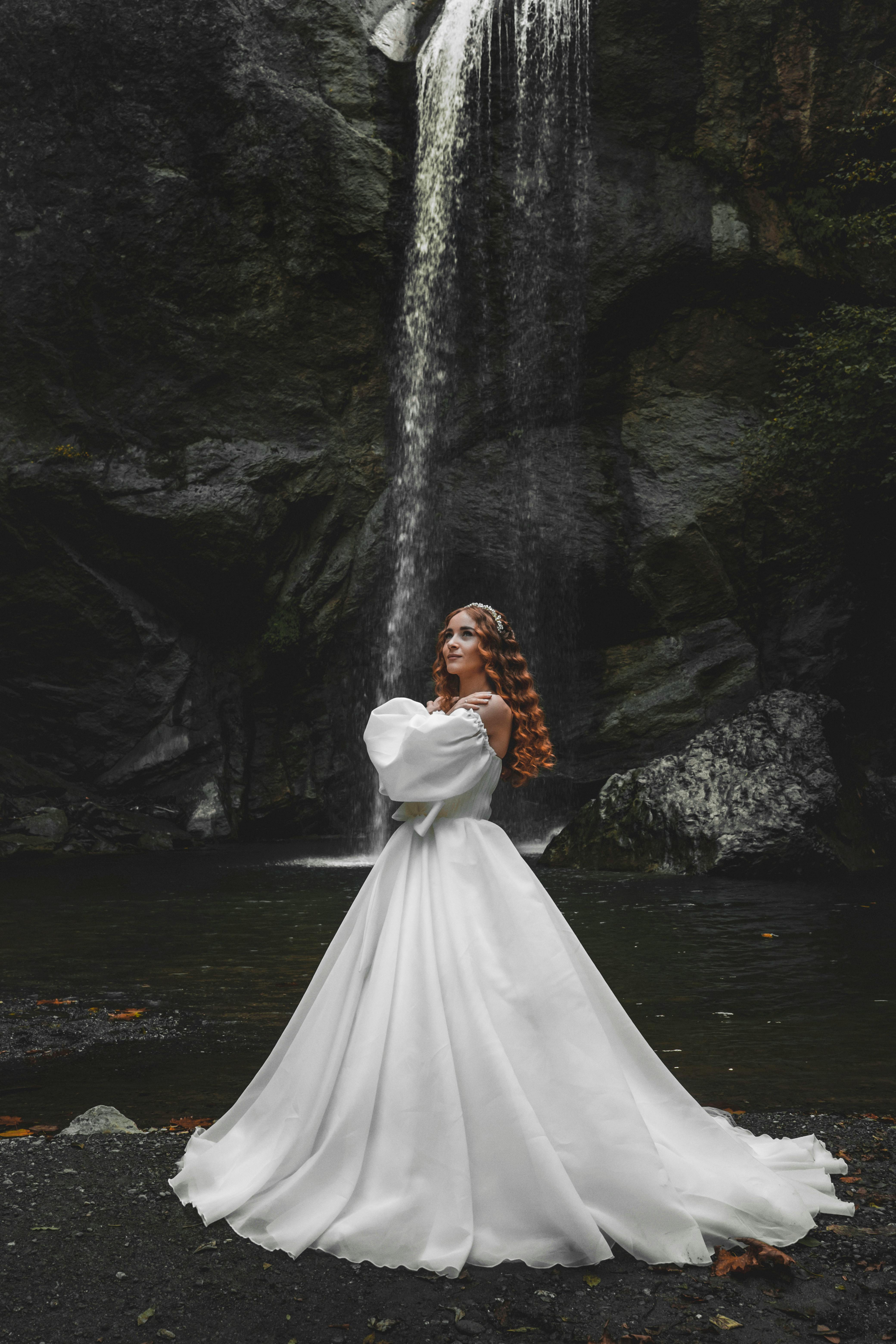 redhead woman in a wedding dress posing by a waterfall