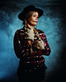 A woman dressed as a scarecrow in a Halloween costume with painted face and hat, set against a moody background.