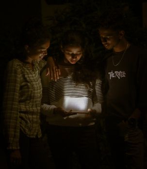 Three young adults using a tablet at night in Madikeri, India, illuminated by its glow.