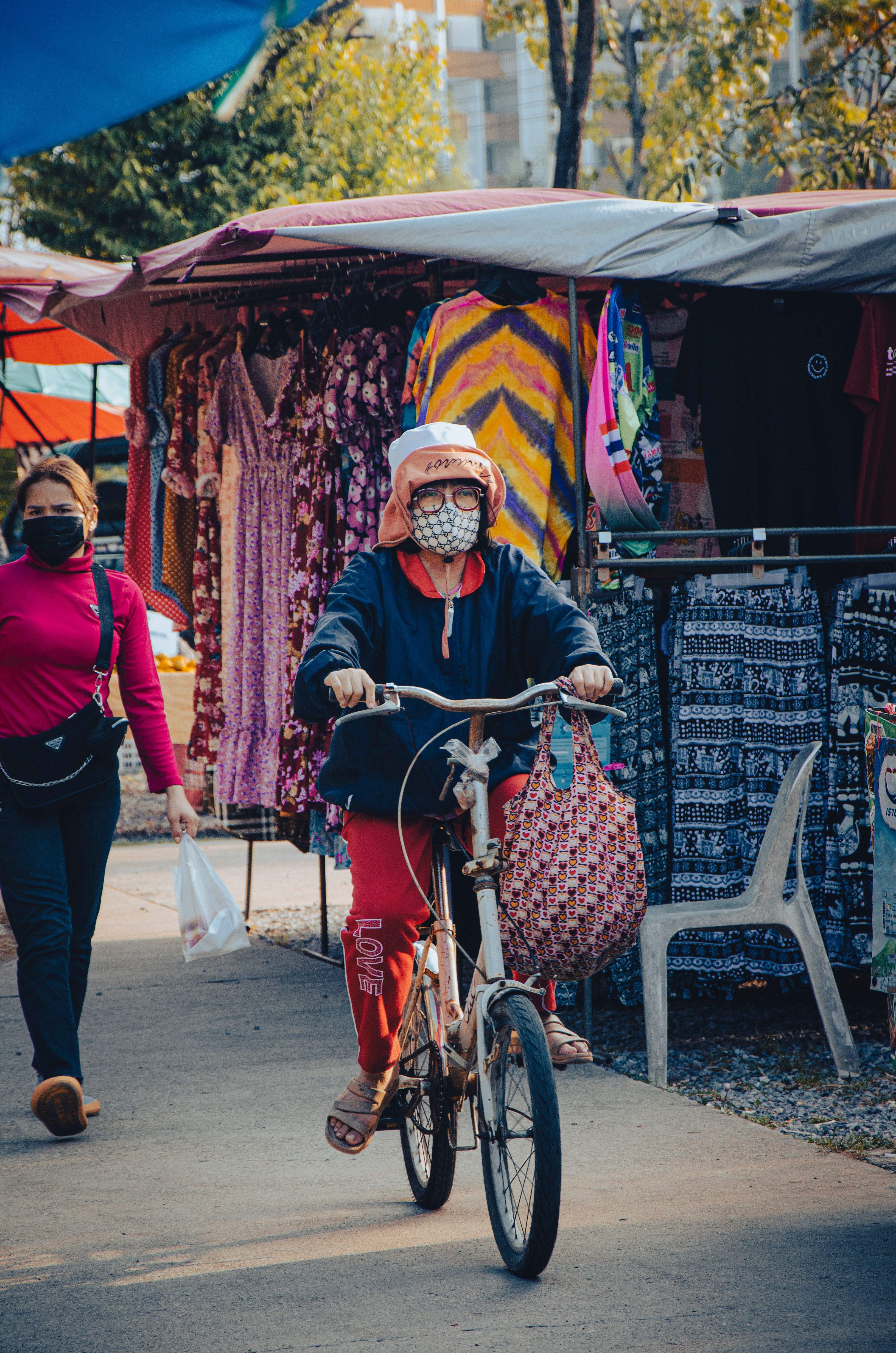 A bustling urban market scene featuring a woman cycling past colorful stalls selling clothes.