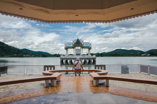 Free A person sitting on a bench overlooking a tranquil lake and ornate pavilion under cloudy skies. Stock Photo