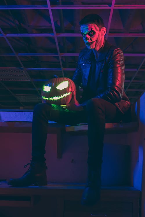 Man in a Halloween Costume and Makeup Sitting and Holding a Carved Pumpkin 