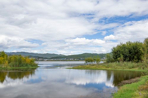 Free A tranquil lake reflecting fluffy white clouds, surrounded by lush greenery and distant hills in summer. Stock Photo