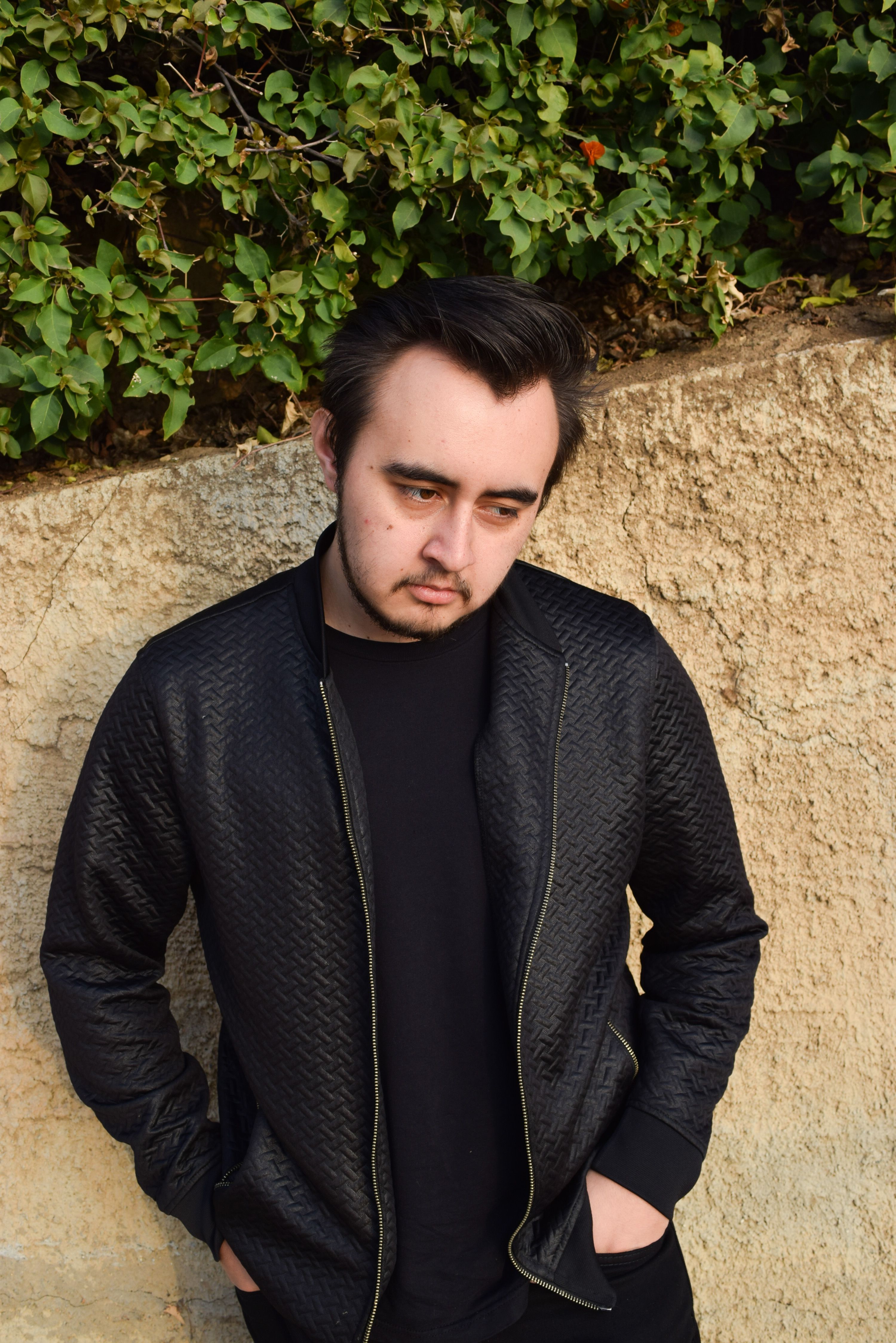 Young man in a black jacket posing by a stone wall with greenery, looking contemplative.