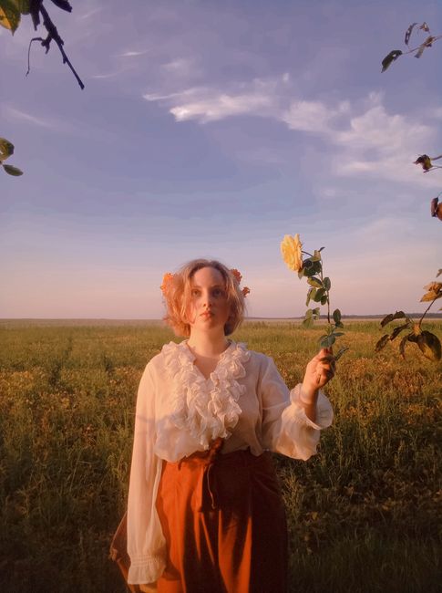 A serene portrait of a woman holding a yellow rose in an open field during a warm sunset.