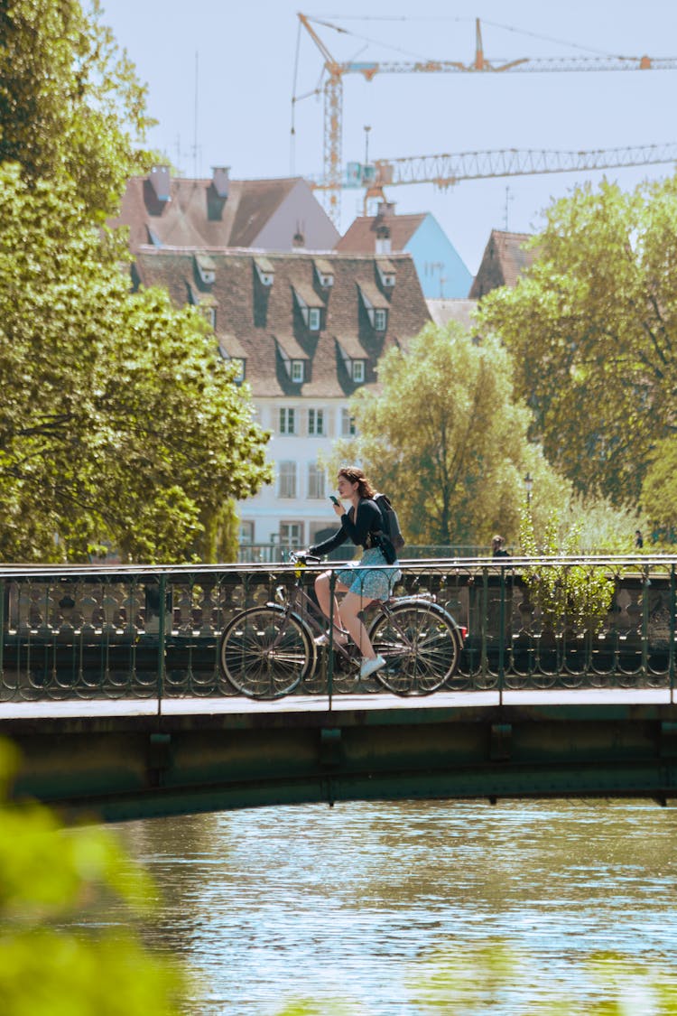 A Woman In Black Long Sleeves Riding A Bike On The Bridge Near The River