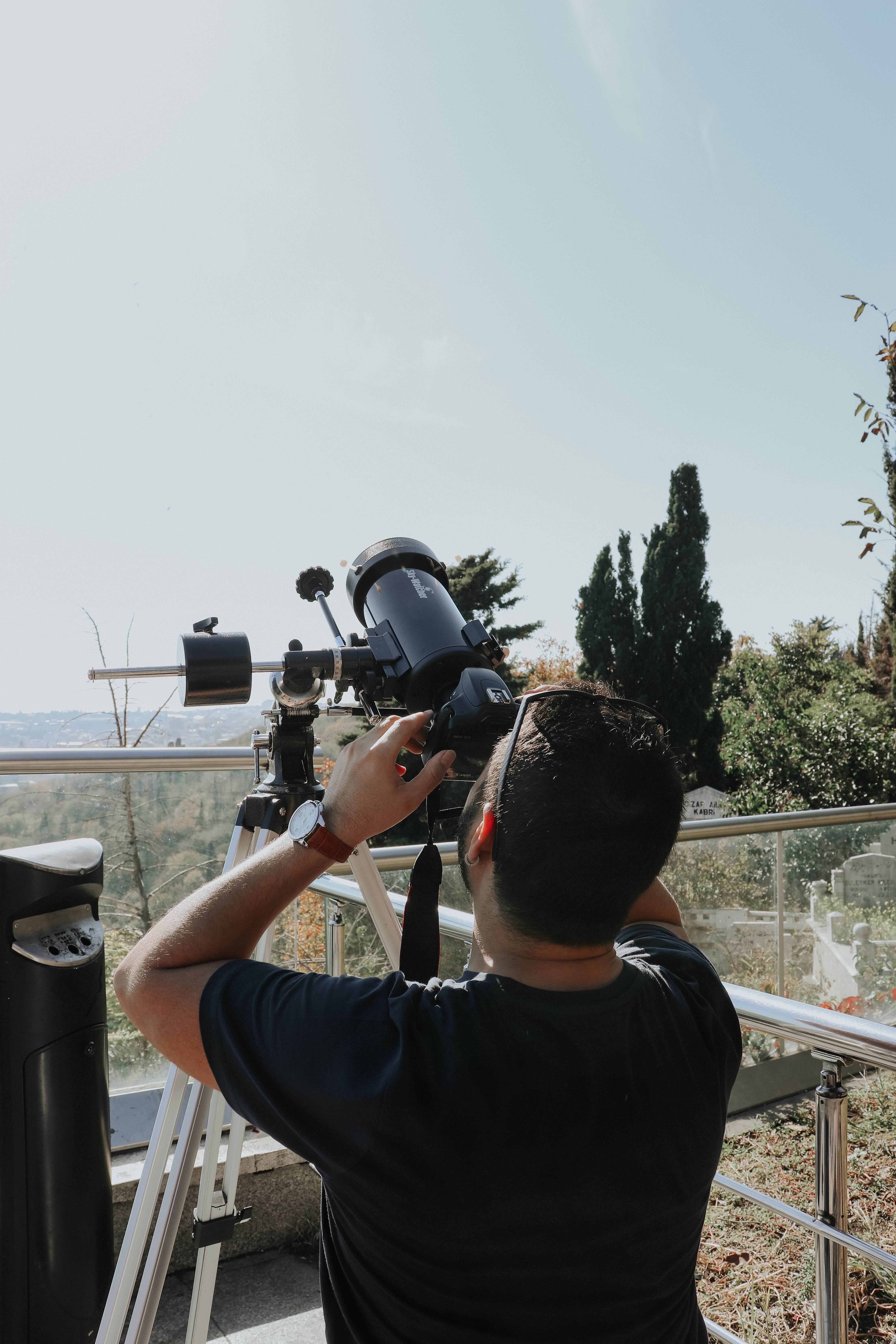 An adult man observes the sky with a telescope outdoors under a clear blue sky.