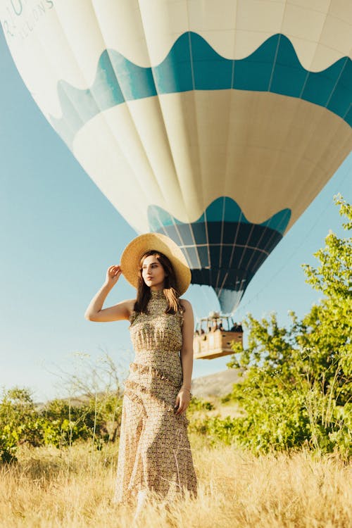 Woman in White and Brown Sleeveless Dress with Sun Hat Standing on Grass Near Hot Air Balloon