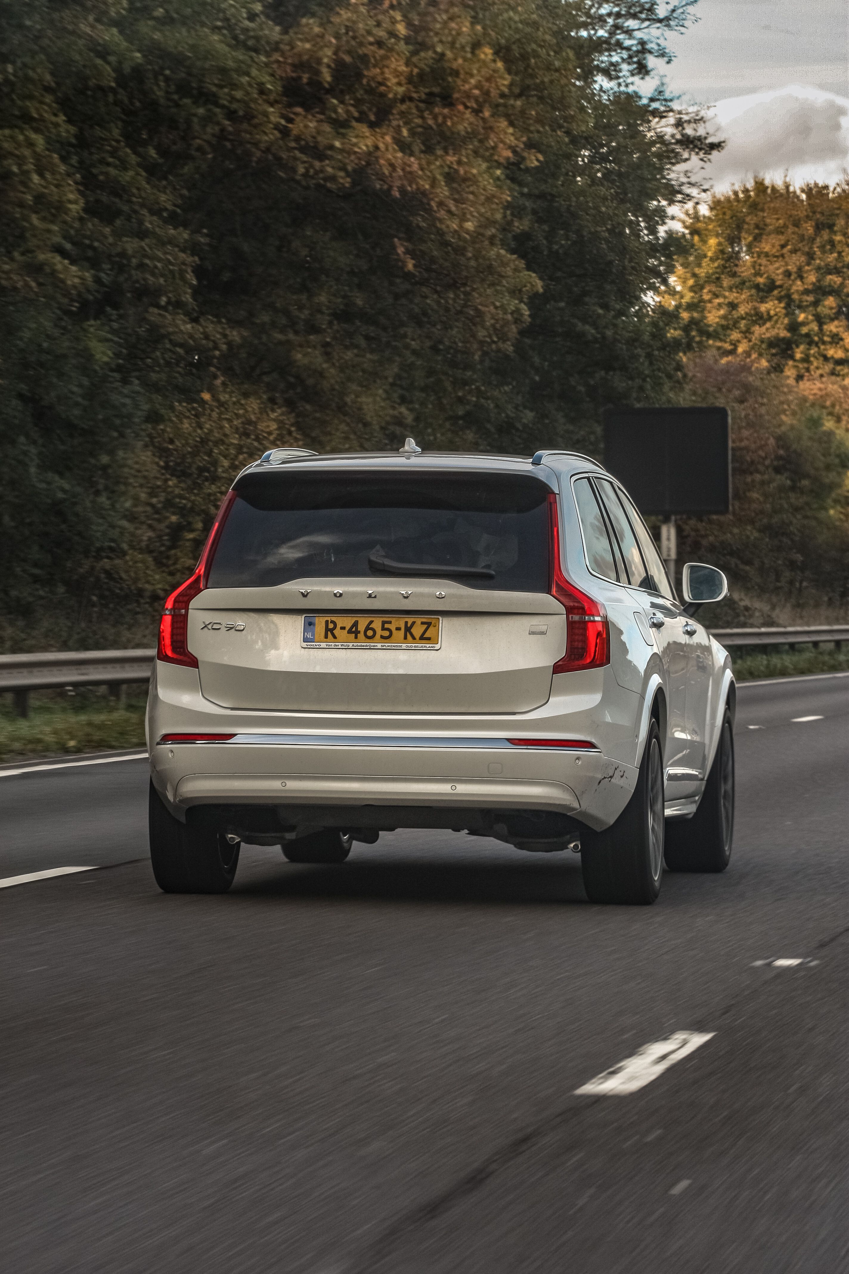 Een witte SUV rijdt op een met bomen omzoomde snelweg tijdens de dag, met zicht van achteren.