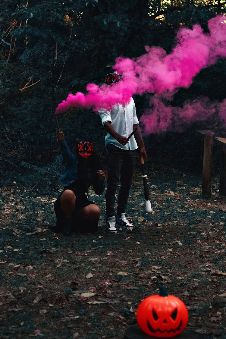 People Wearing Masks Playing With Pink Smoke In A Dark Park At Halloween