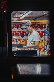 A food truck vendor wearing a face mask prepares food inside a vibrant, colorful setting.