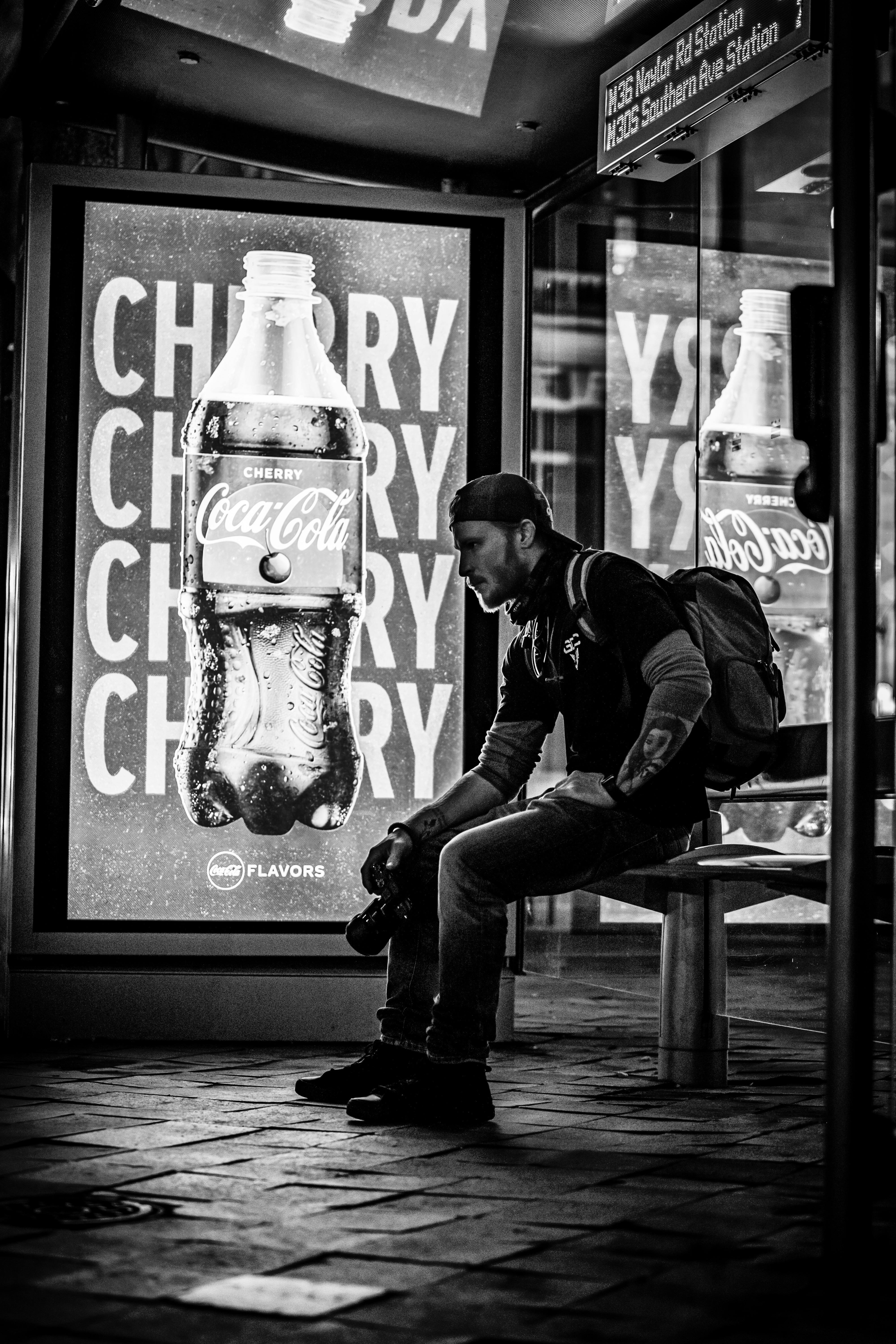 Black and white image of a photographer resting at a city bus stop with a cherry cola ad.