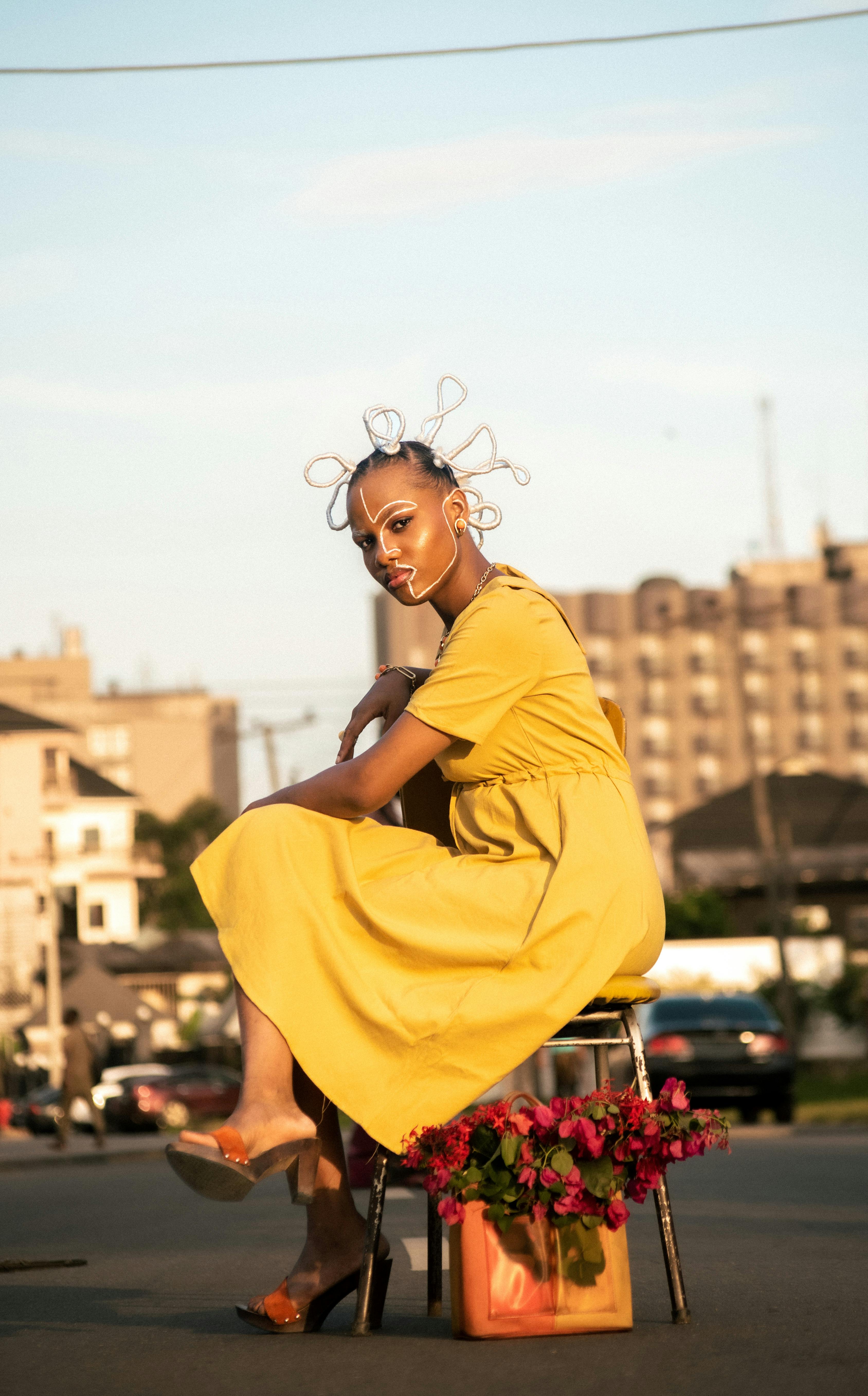 A fashionable woman in a unique hairstyle and yellow dress poses by flowers on a street in Port Harcourt, Nigeria.