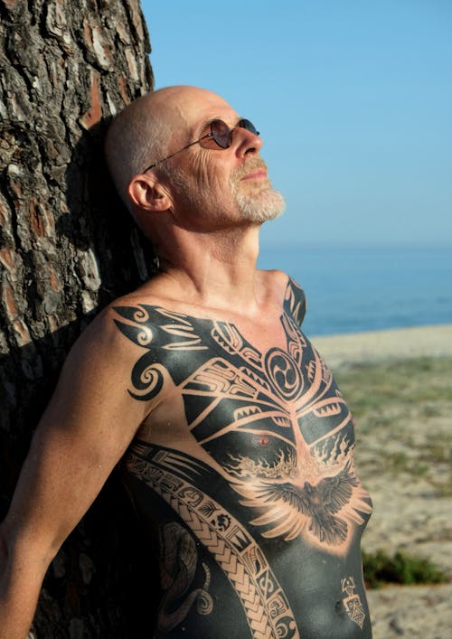 Man with Body Tattoo Leaning on Tree Trunk