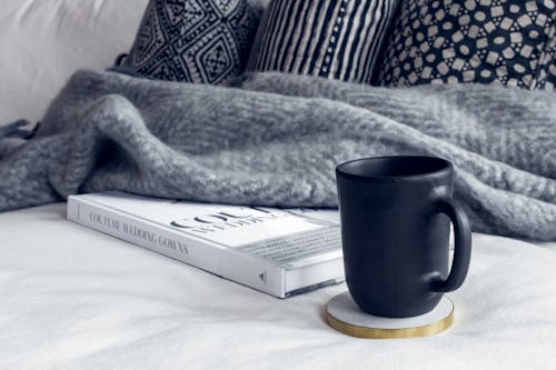 Caneca De Cerâmica Preta Em Porta Copos Redondo Branco E Bege Em Tecido Branco Ao Lado Do Livro